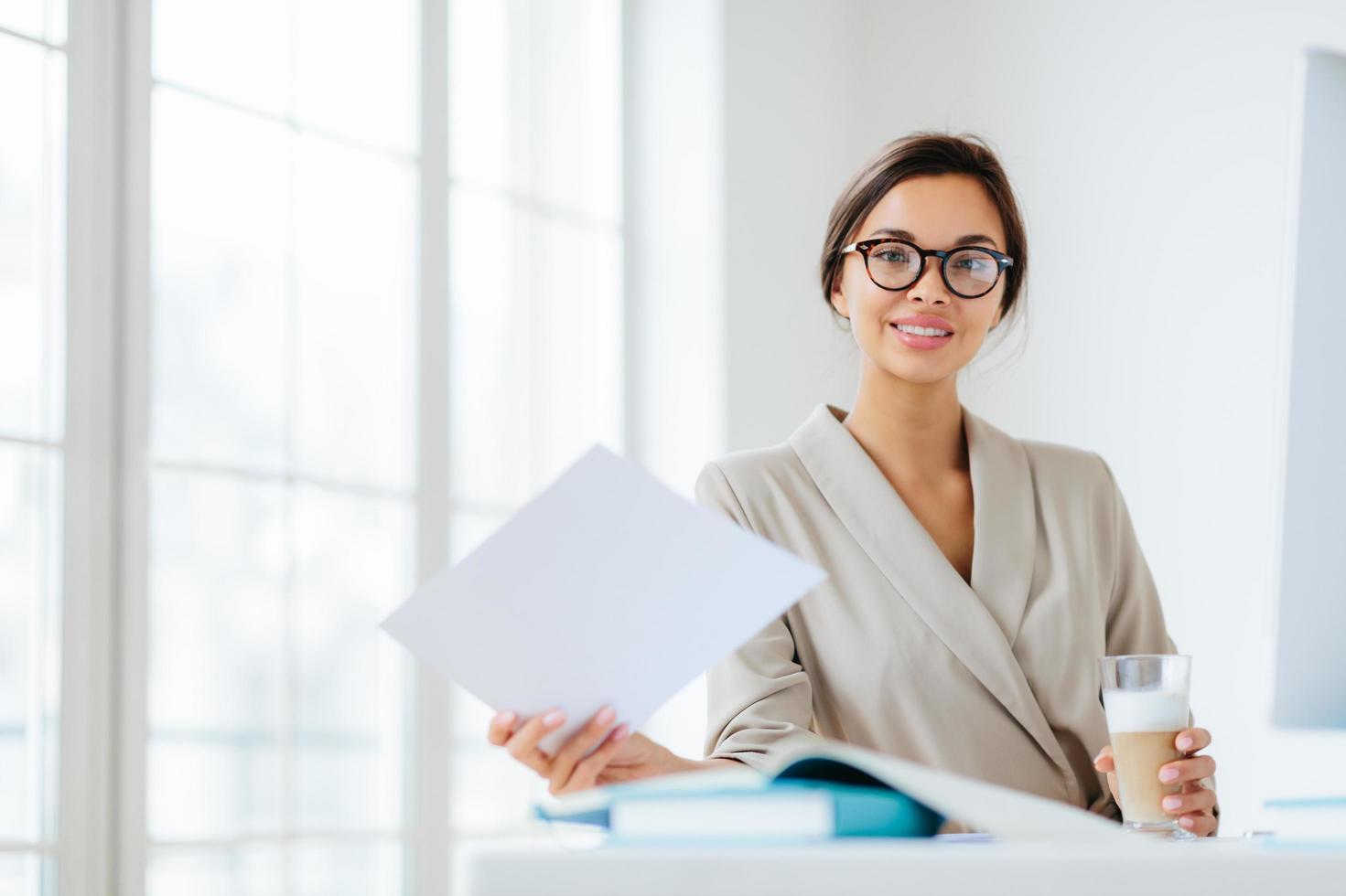 Photo of delighted dark haired European woman in transparent glasses, works with documents, reads information data, dressed in elegant clothing, drinks milkshake sits at desktop in office makes report