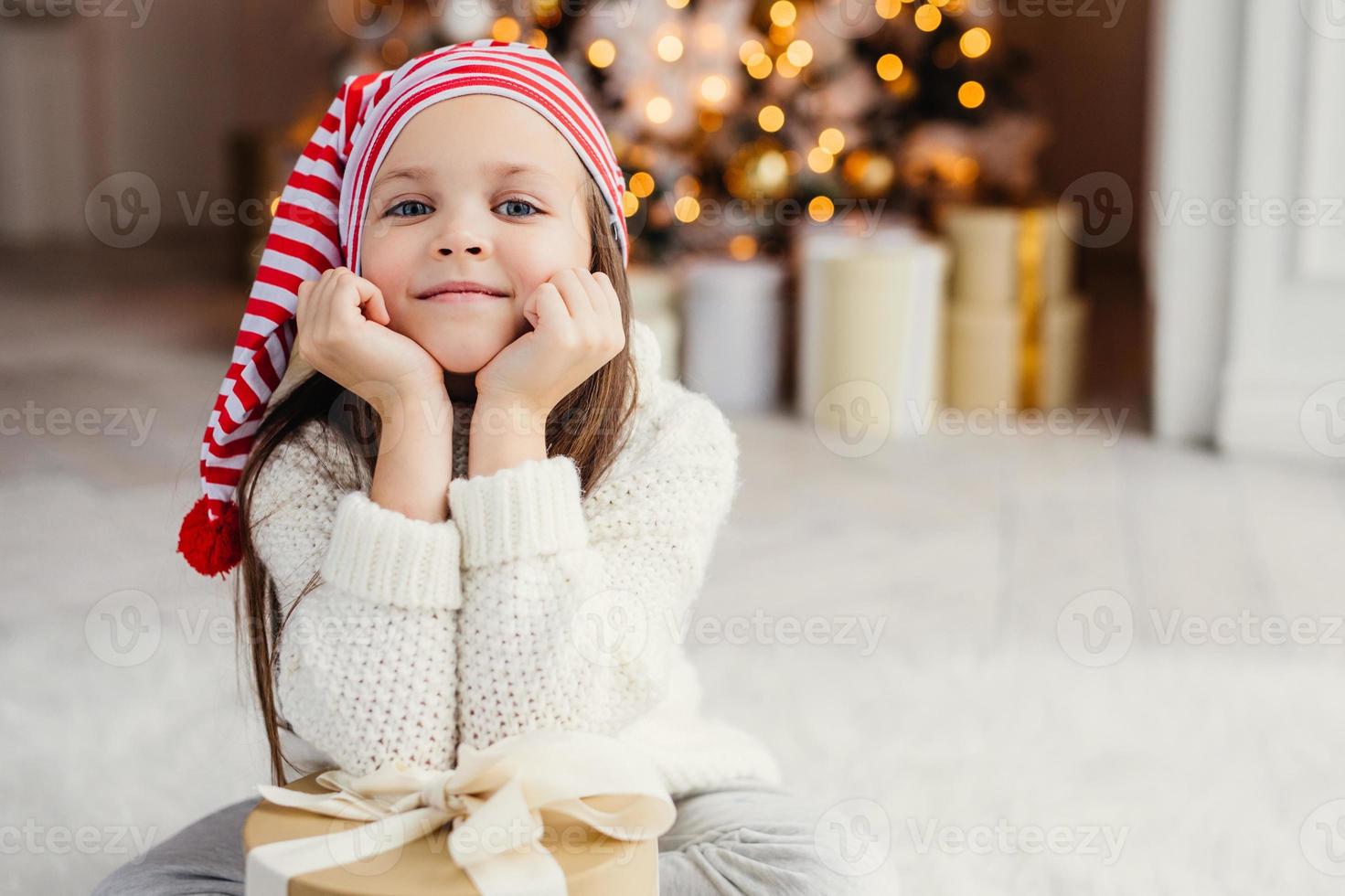 Horizontal portrait of adorable little child, leans on hands with present box, sits against decorated Christmas tree. Blue eyed small kid in knitted white sweater poses at camera. Holidays concept photo