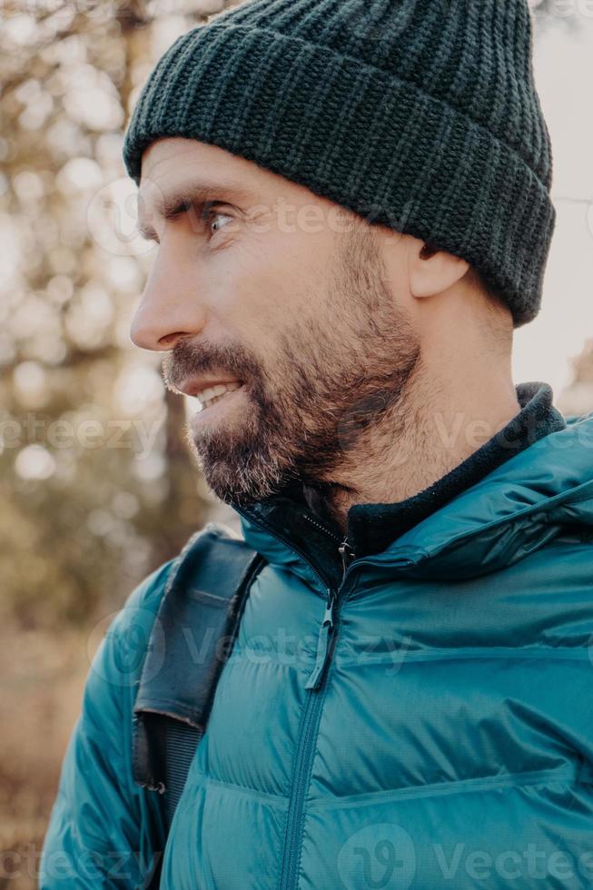 Sideways shot of unshaven man with dark thick beard, wears warm har and anorak, looks pensively aside, poses outside, dreams about something has outdoor stroll in unknown place alone, active lifestyle photo