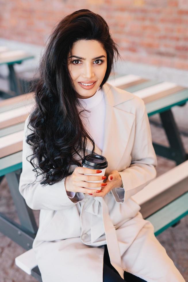 Portrait of adorable woman with dark long wavy hair, bright eyes and full lips wearing white coat sitting at bench in outdoor cafe drinking takeaway coffee posing in camera smiling pleasantly photo