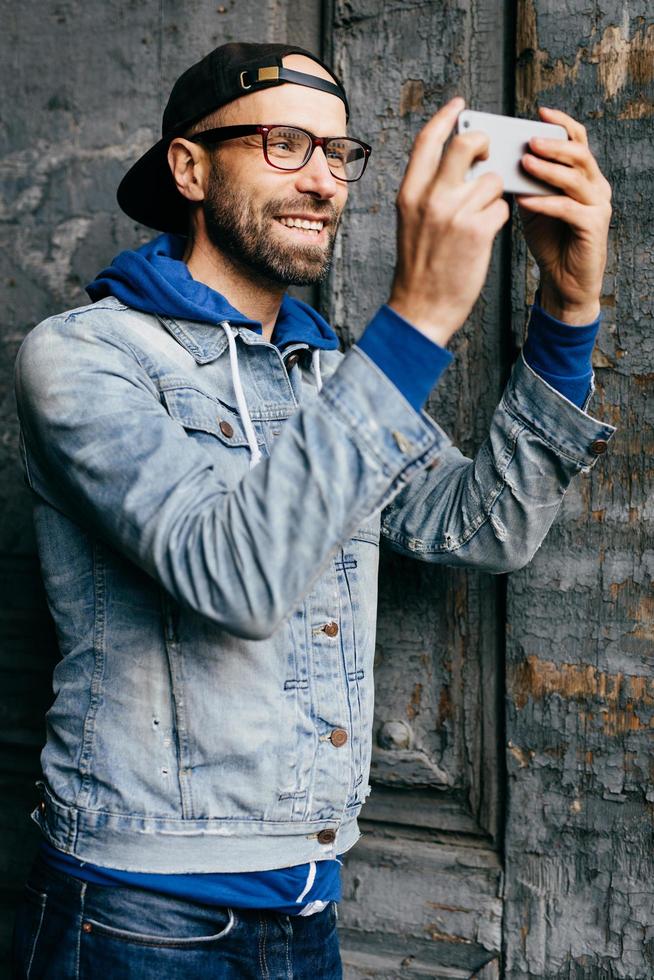 Excited male with beard and moustache wearing trendy denim shirt with hook and jeans looking happily into camera of his smartphone making selfie. Wonderful hipster man using modern device isolated photo