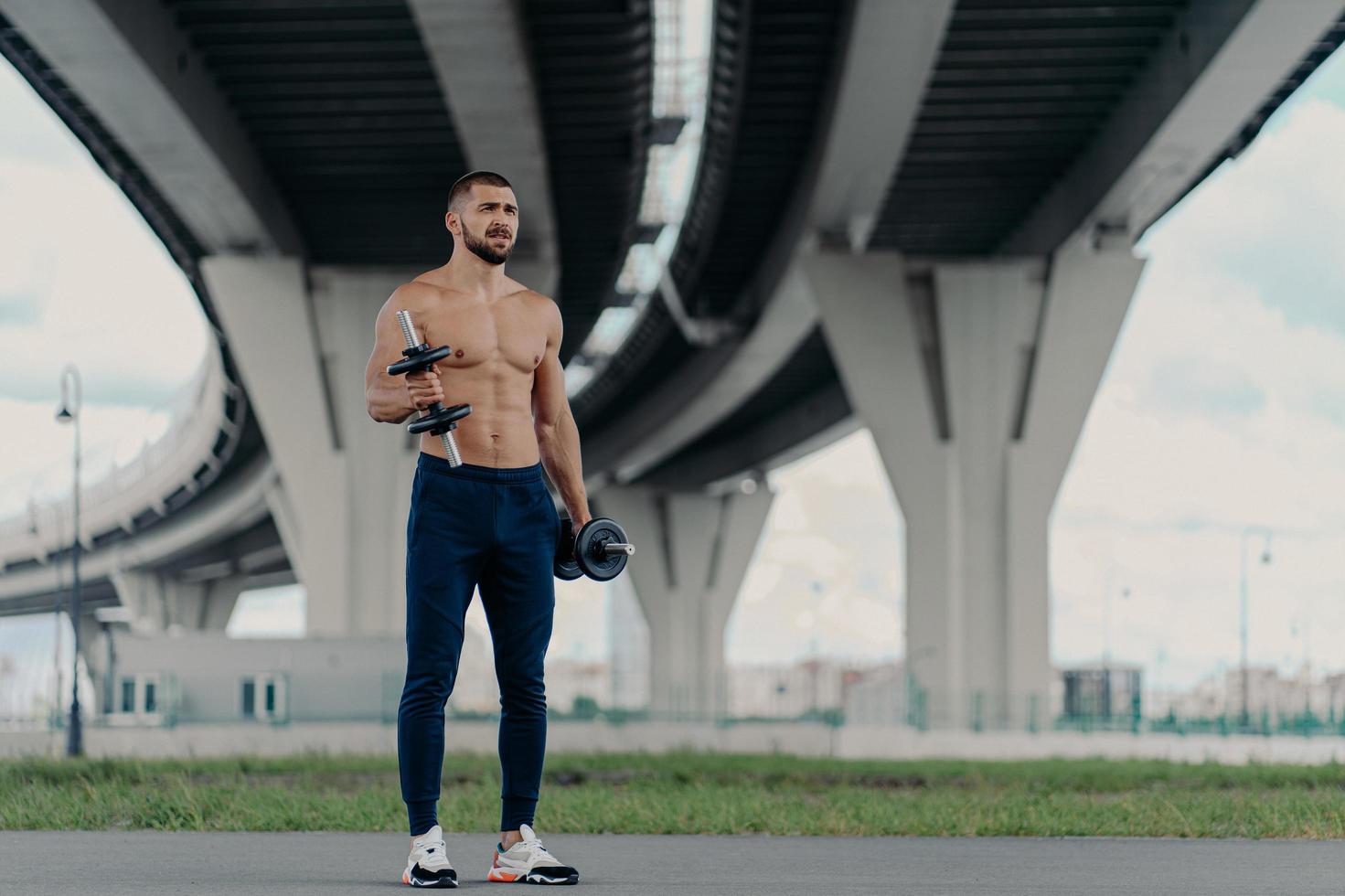 Muscular athletic bearded bodybuilder raises dumbbells works on biceps has hard workout near bridge dressed in sport trousers and sneakers, being motivated, enjoys sport. Healthy lifestyle concept photo