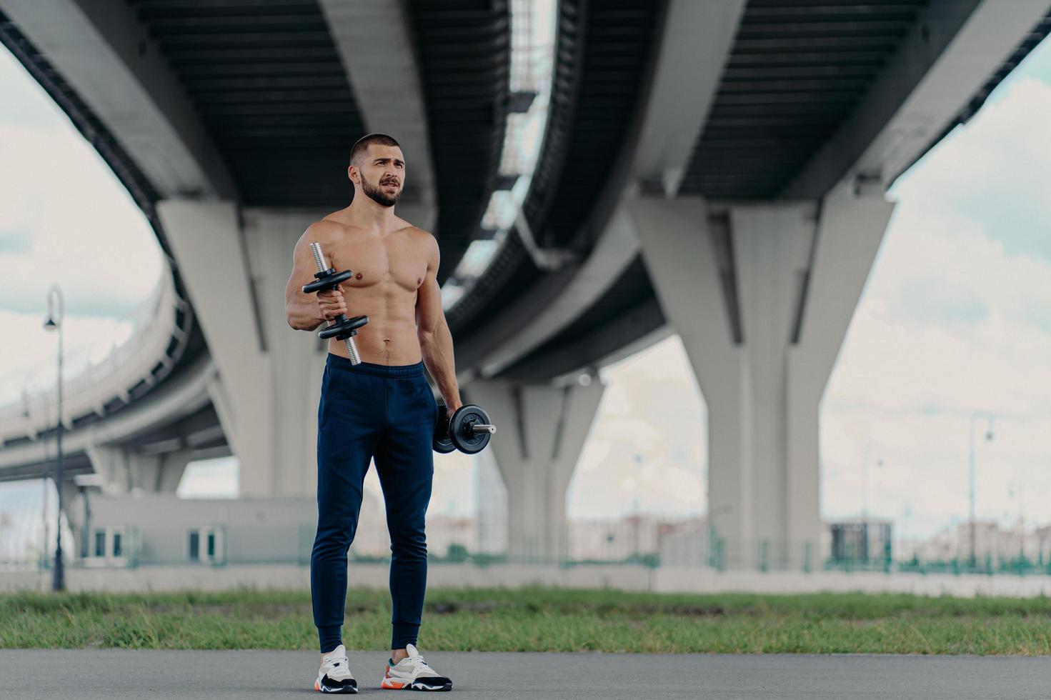 musculoso culturista barbudo atlético levanta pesas trabaja en bíceps tiene entrenamiento duro cerca del puente vestido con pantalones deportivos y zapatillas de deporte, está motivado, disfruta del deporte. concepto de estilo de vida saludable foto