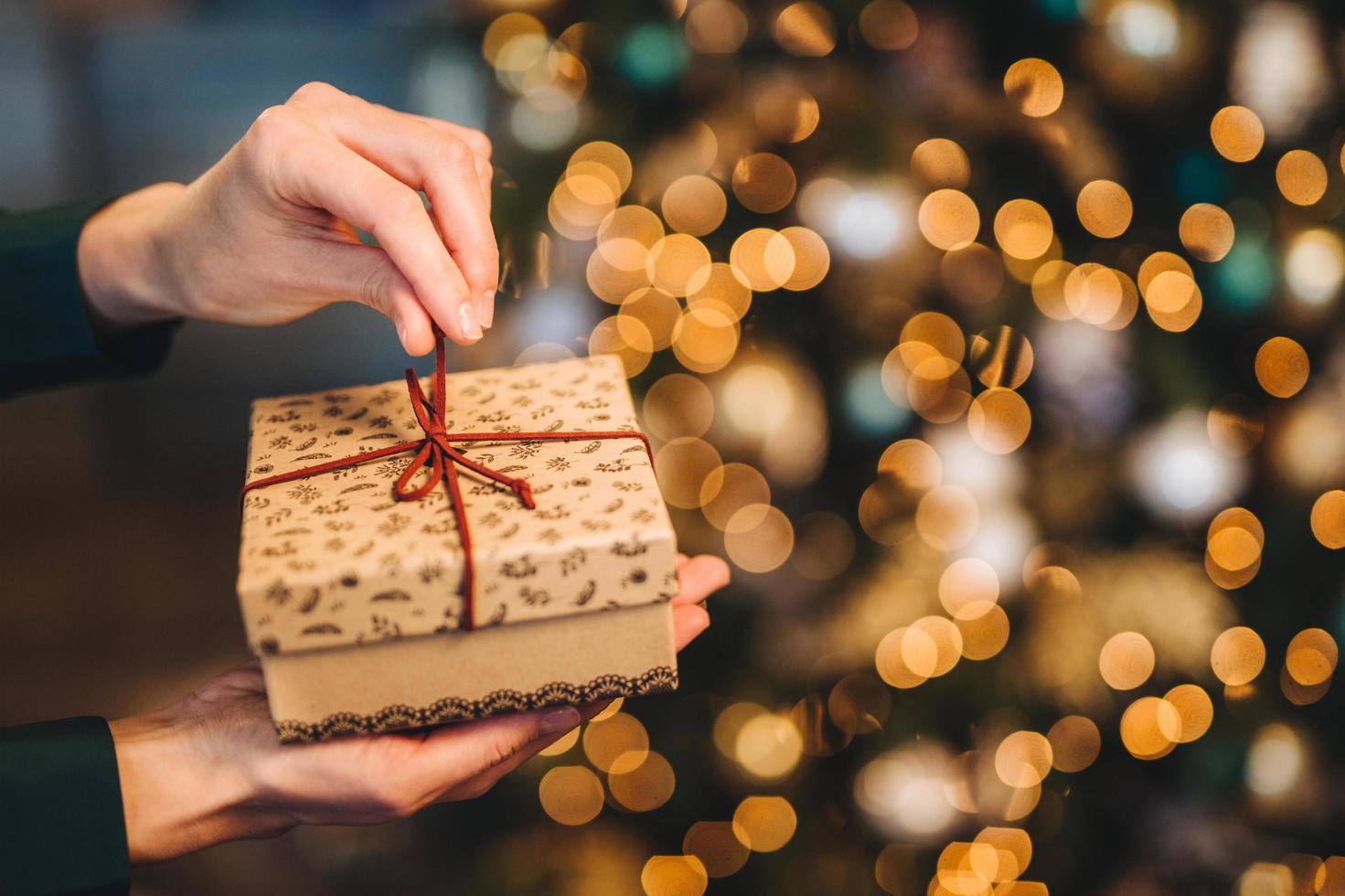 Unrecognizable woman wrappes gift box over Christmas tree with shining lights and garlands. Decorated present box in womana hands. Holidays and celebration concept. photo