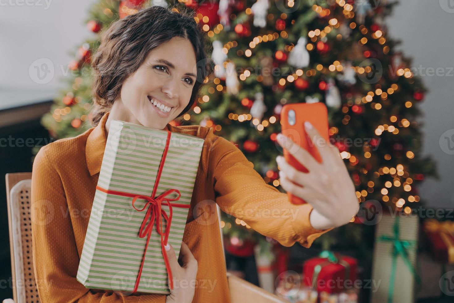 Cheerful Italian female sitting near beautiful Christmas tree at home and taking selfie with gift photo