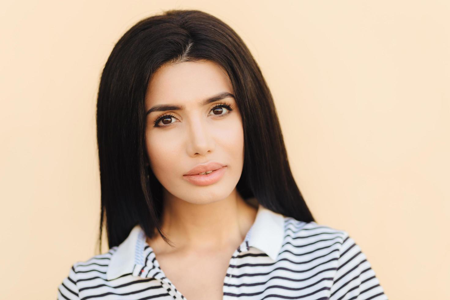 Serious beautiful brunette female looks confidently into camera, has long straight hair, listens attentively interlocutor, wears casual striped t shirt, isolated on light background. People and beauty photo