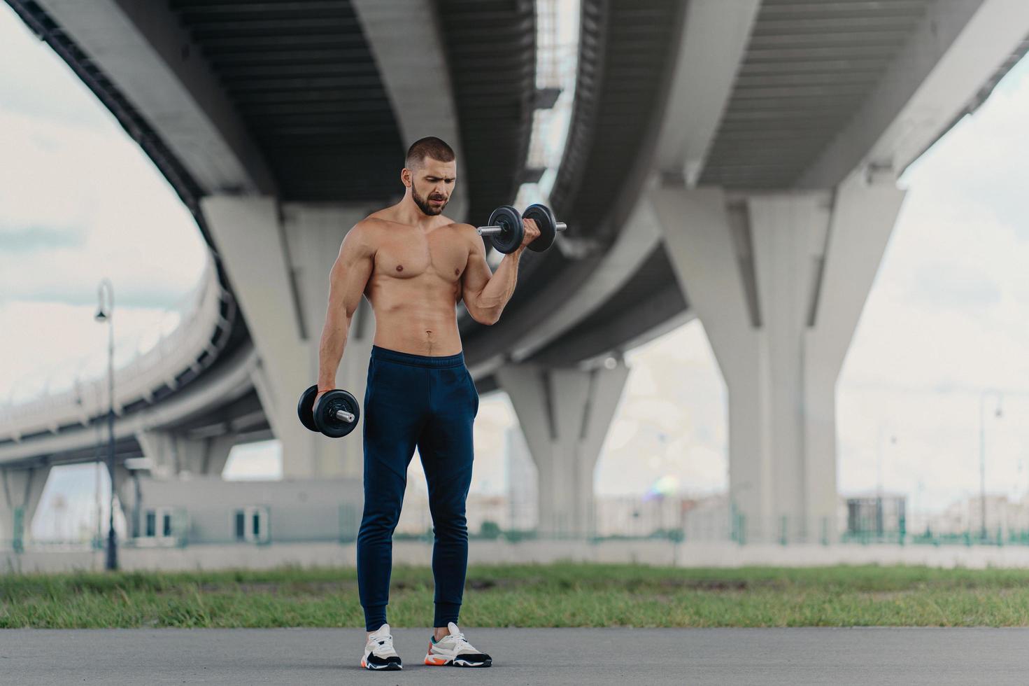 un culturista fuerte y saludable levanta pesas pesadas, hace ejercicio al aire libre, demuestra su fuerza, tiene bíceps, se para debajo del puente y mantiene una buena forma física. joven deportista trabaja en los músculos foto