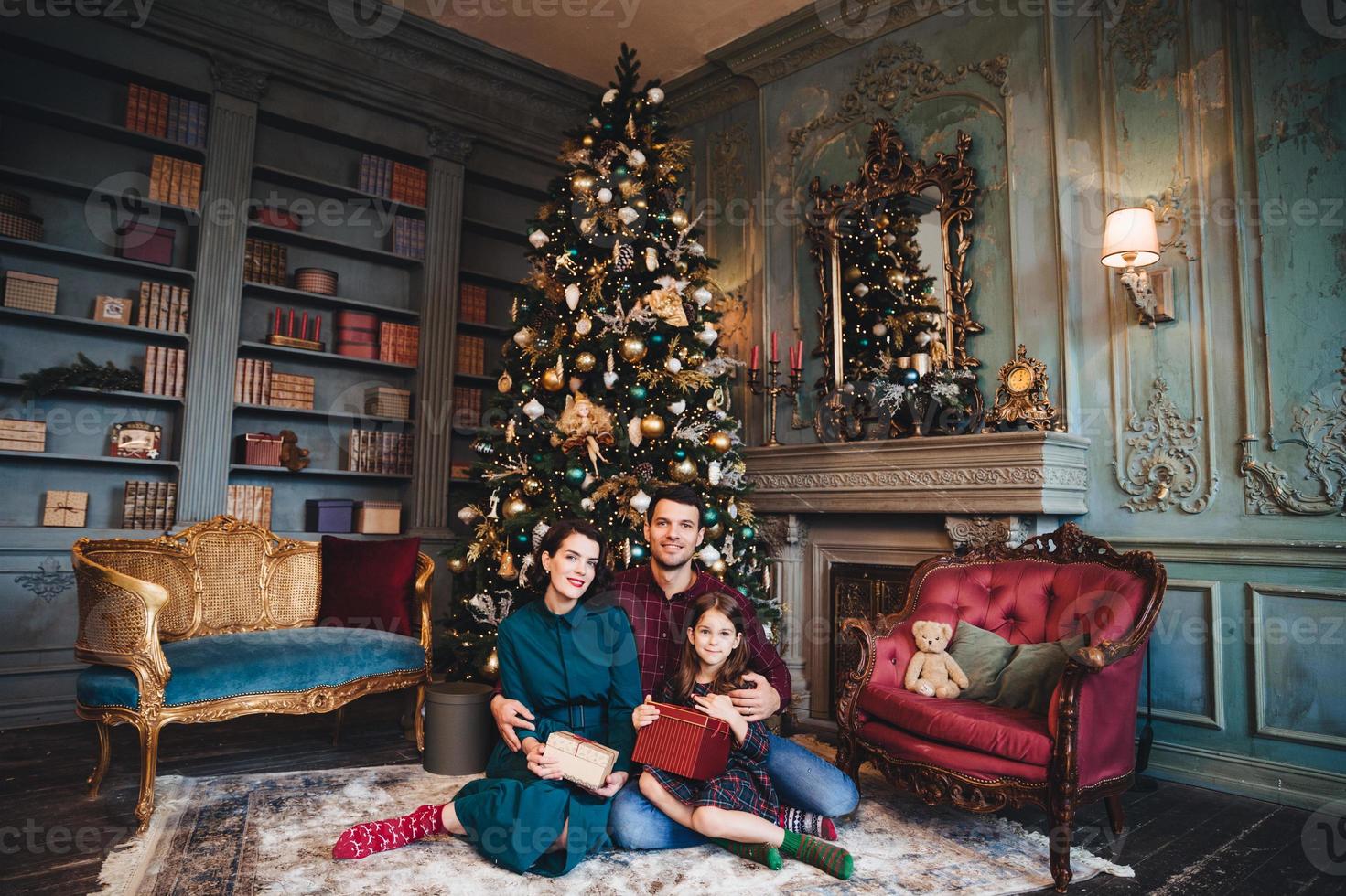 Friendly family sits near decorated Christams tree, embrace each other, hold packed presents, enjoy majestic atmospere. Father, mother and daughter near New Year tree in living room. Festive concept photo