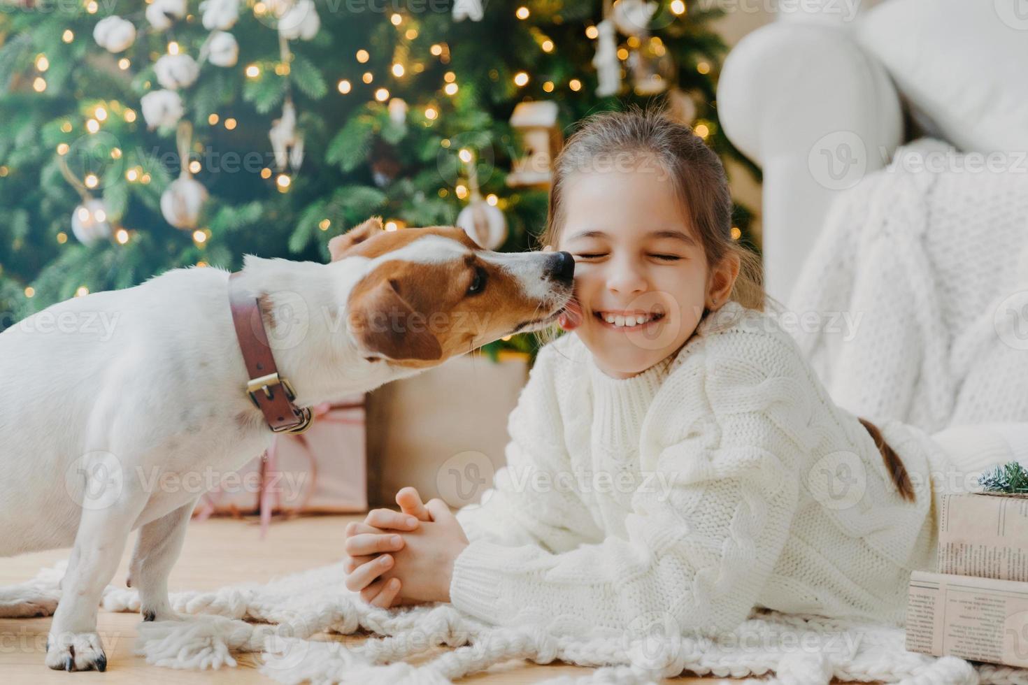 Lovely puppy licks chillds face have fun together pose on floor in cozy room against decorated fir tree, present boxes. Happy New Year concept. Beginning of winter holidays. Christmas preparation photo