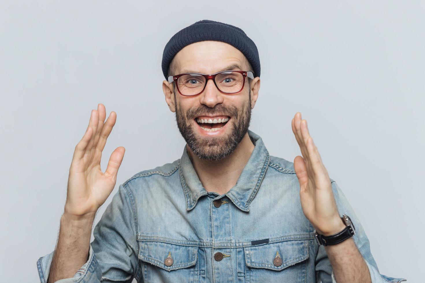 Handsome happy man raises hands with excitment, has overjoyed expression, looks with delighted face, has thick beard and mustache. Glad man in eyewear poses against white background, has fun photo