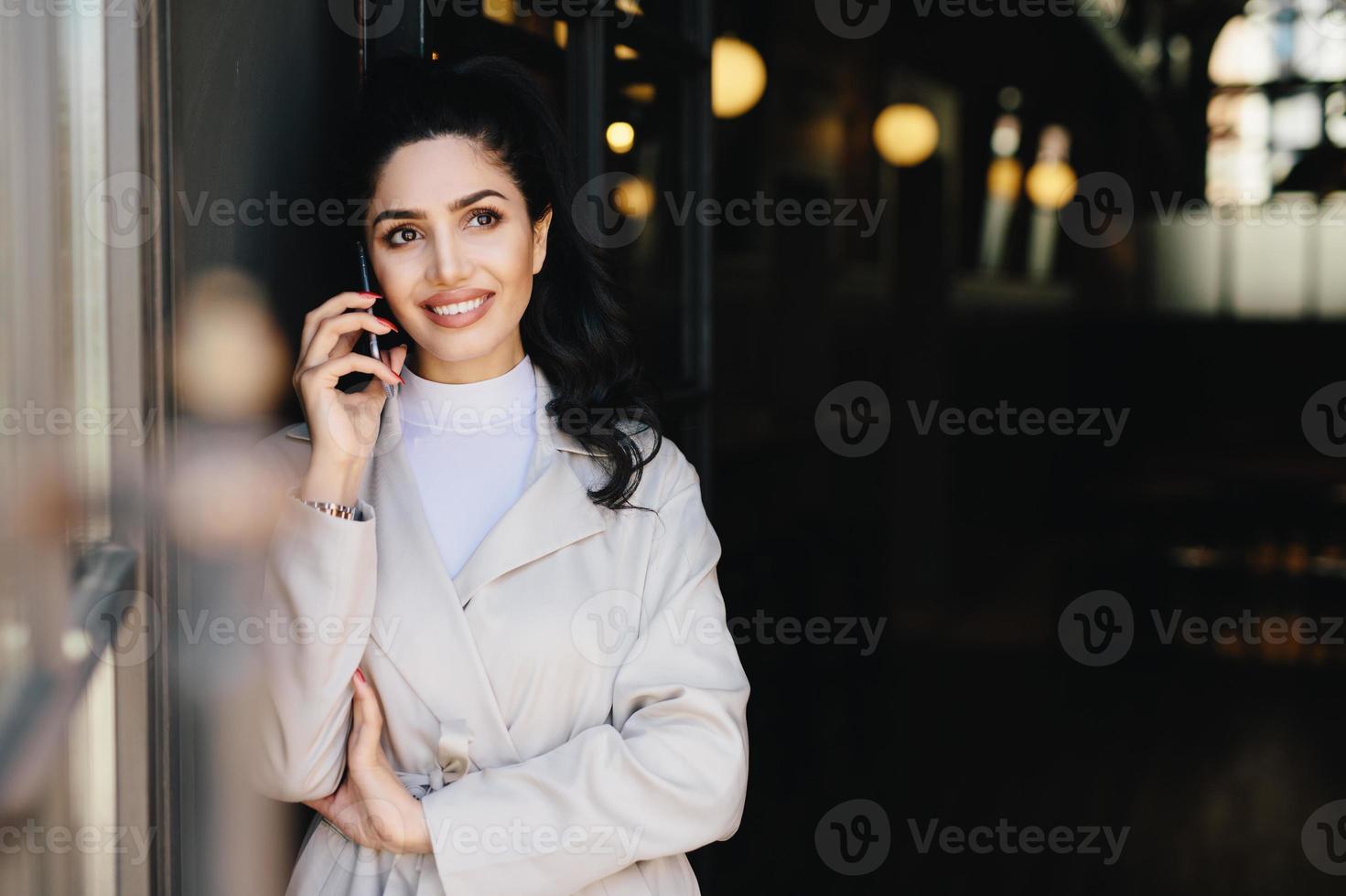 Elegant brunette businesswoman with dark eyes and full lips dressed in white clothes communicating over telephone having charming smile looking up with thoughtful expression. Technology concept photo
