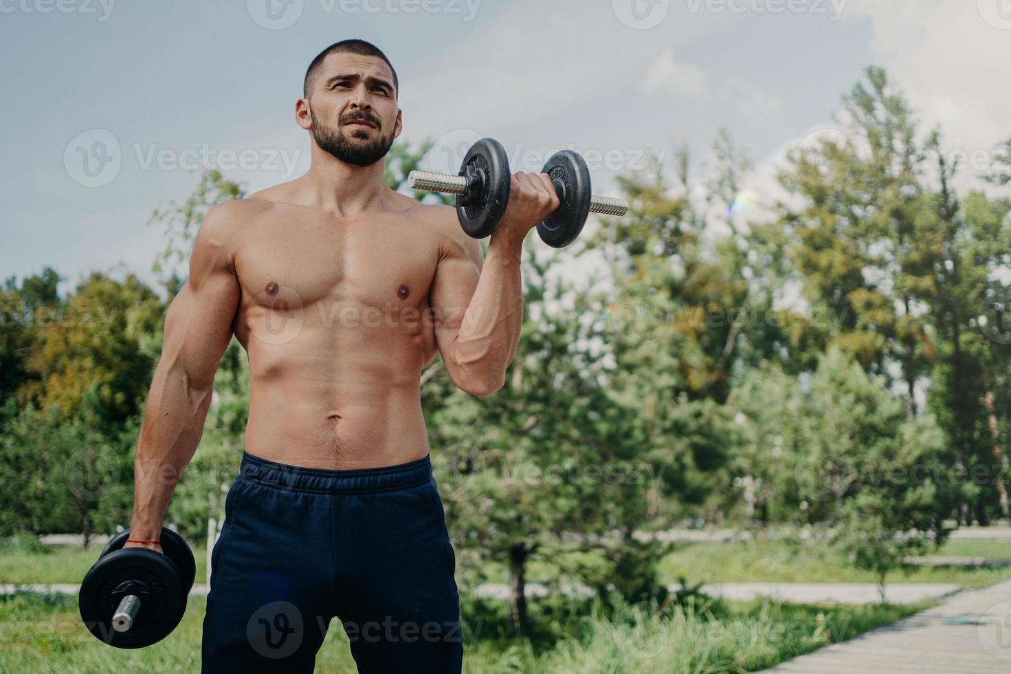 tiro horizontal de un hombre europeo sin afeitar en topless fitness tiene un cuerpo musculoso, levanta pesas, usa pantalones cortos, demuestra brazos fuertes, hace ejercicio al aire libre en el parque. deportista hace levantamiento de pesas foto