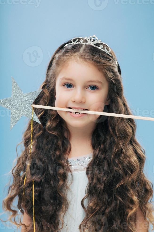 Photo of attractive woman kid with pleasant appearance, has long dark hair, holds magic wand in mouth, poses over blue background, dressed in princess dress. Vertical image. Childhood concept