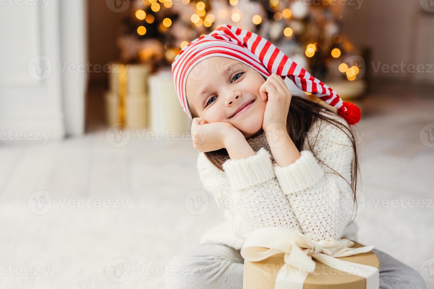 un hermoso niño pequeño posa en la sala de estar, se apoya en el regalo actual, tiene una expresión feliz, se alegra de recibir la sorpresa de los padres, pasa las vacaciones en un círculo familiar. Feliz navidad y próspero año nuevo foto