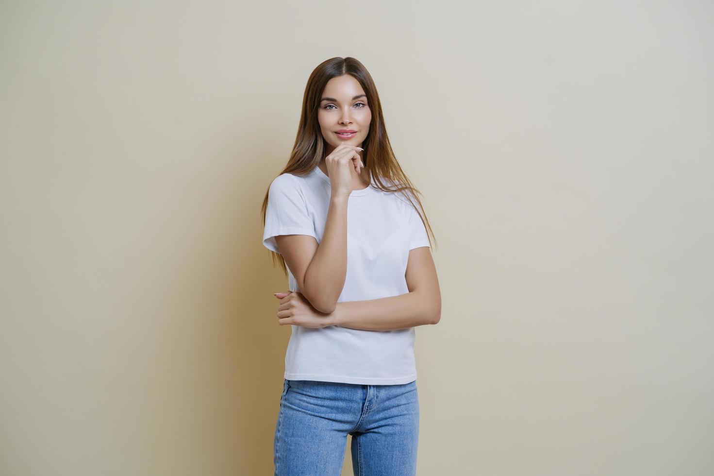 Photo of brunette young European woman holds chin, wears white t shirt and jeans, enjoys conversation with her mates, isolated over brown background. People, lifestyle, face expressions concept