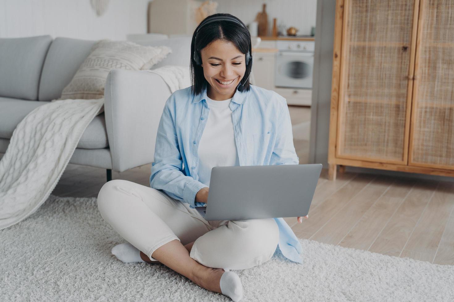 Smiling woman in headset working on laptop from home learning online sitting on the floor. Elearning photo