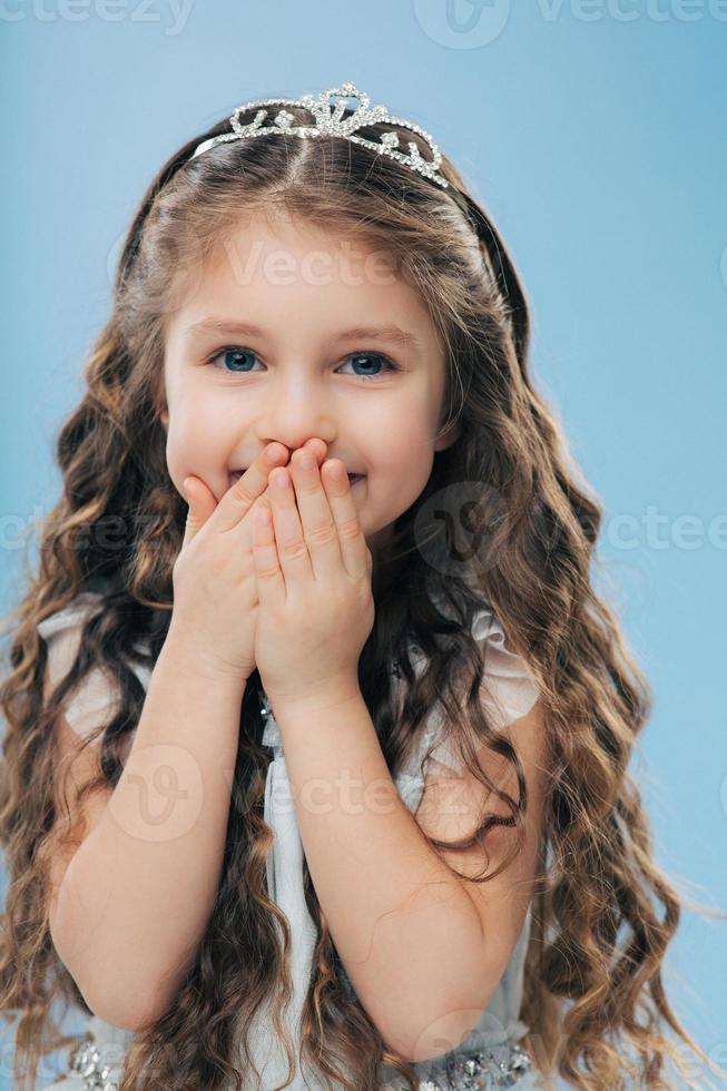 Image of positive beautiful smiling child keeps both hands on mouth, has long thick hair, blue eyes, wears crown, poses against blue background, expresses happiness. Children and satisfaction photo