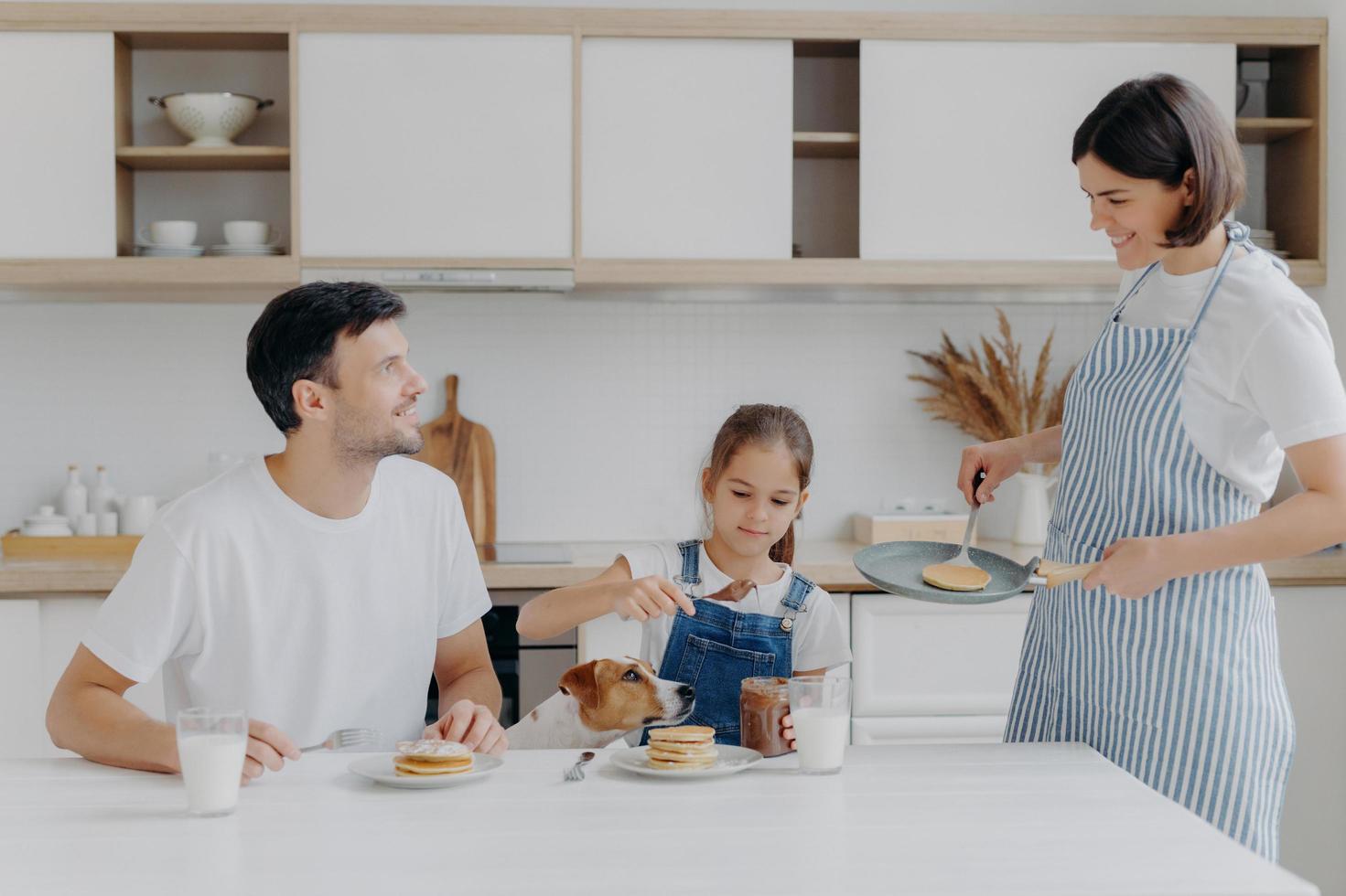 feliz tiempo en familia y concepto de desayuno. la alegre esposa y la madre preparan deliciosos panqueques para los miembros de la familia, el padre, la hija y el perro disfrutan comiendo y degustando el postre en casa, agregan chocolate foto