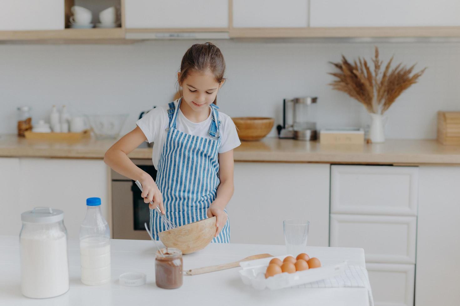 Cute small girl wears striped apron, whisks ingredients in bowl, prepares dough, teaches to cook, stands at white table with eggs, milk, flour against kitchen background. Childern and cooking concept photo