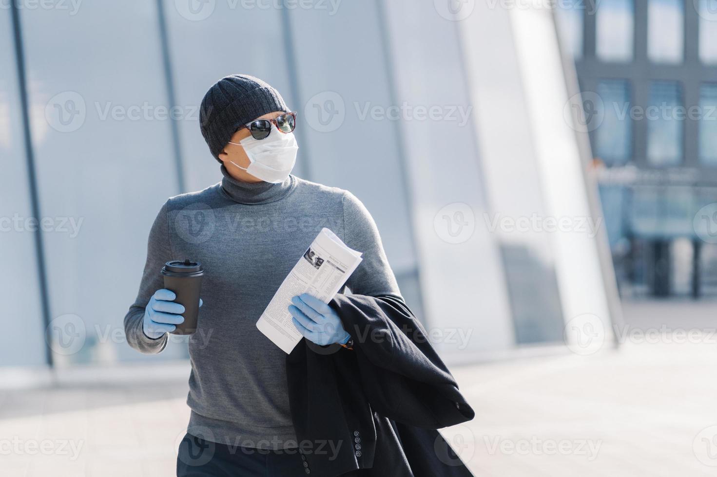 Shot of man walks outdoor, protects from flu, coronavirus spreading, drinks coffee, looks aside with pensive expression, reads newspaper, follows quarantine rules. 2019-ncov. Pandemic virus. photo