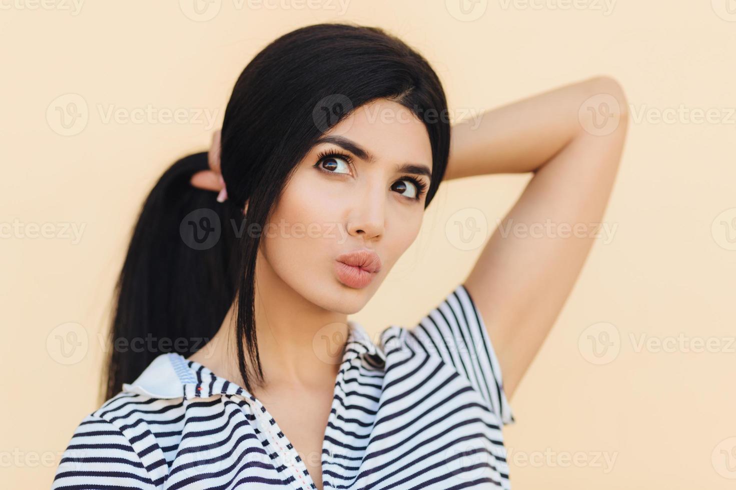 Cute young woman has dreamy expression, keeps lips round, has make up, makes hair in pony tail, dreams about something, looks thoughtfully, isolated over beige background. Youth and beauty concept photo