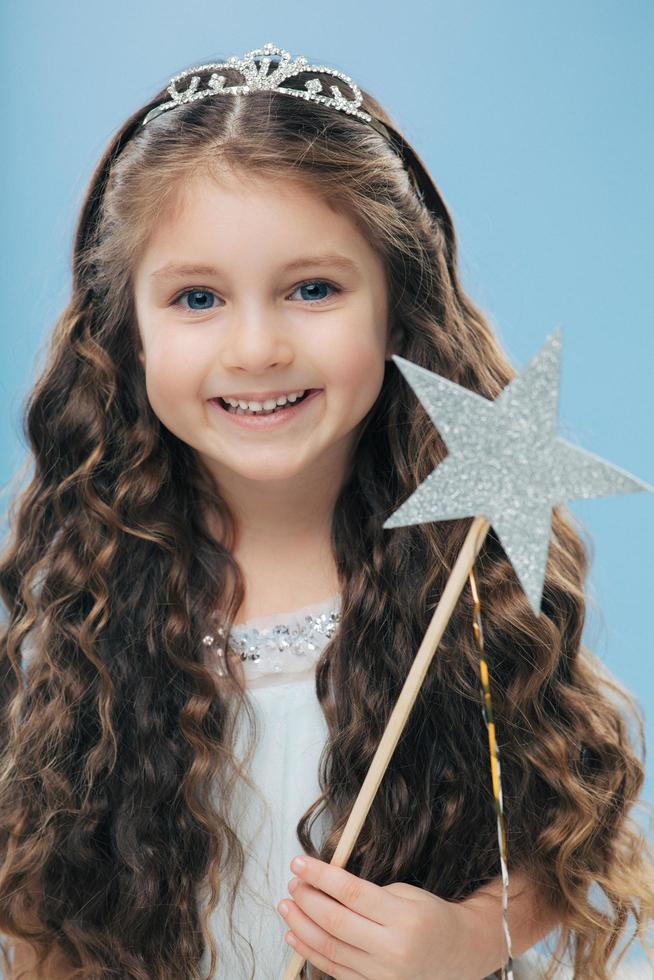 Cute little girl with positive expression, holds wand, has curly dark hair, carries wand, believes in fairy tale, isolated over blue background, wishes dreams come true. Children and magic concept photo