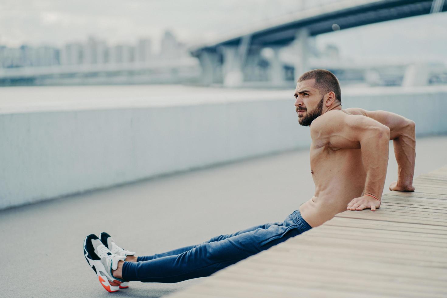 guapo deportista entrena los músculos y hace ejercicios de flexiones inversas se calienta al aire libre antes de correr, se concentra a un lado, tiene un cuerpo musculoso, lleva un estilo de vida activo y saludable, tiene un torso musculoso desnudo foto