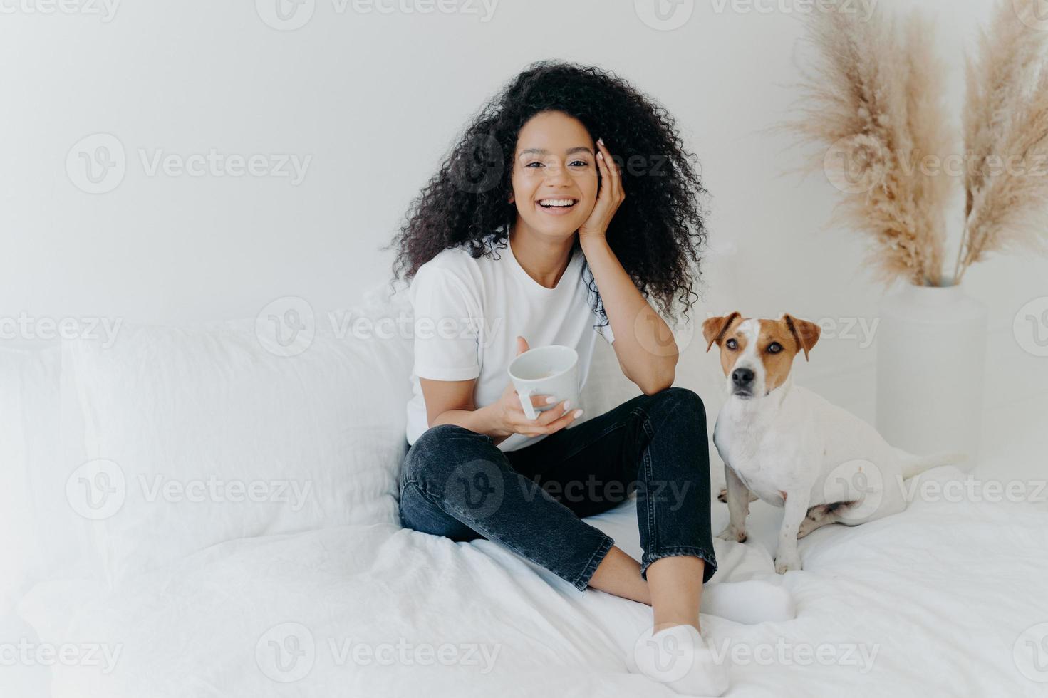 Happy attractive ethnic woman with curly hair wears white t shirt, jeans and socks, smiles pleasantly, drinks tea in comfortable bed, poses with dog, have lazy weekend. People, rest, animals concept photo