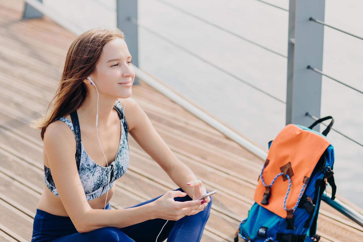 Sideways shot of dreamy pleased female model looks thoughtfully aside, listens favourite music, holds modern cell phone, chooses track in playlist, connected to high speed internet, poses outdoor photo