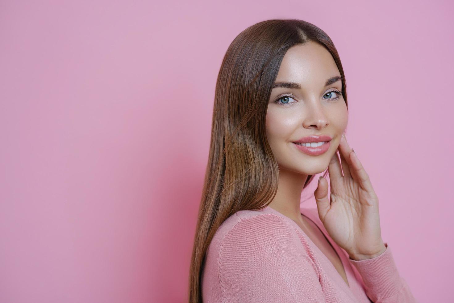 Photo of beautiful brown haired Caucasian woman keeps hand on cheek, smiles gently and looks directly at camera, has blue eyes, wears minimal makeup, stands in profile against pink background