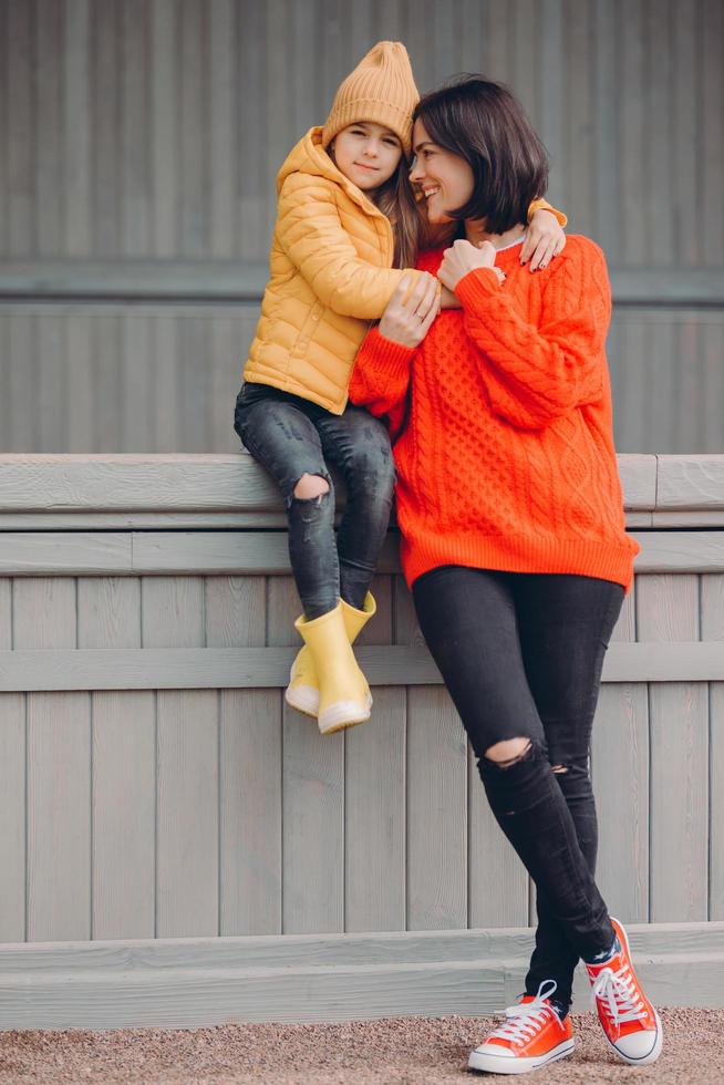 Photo of affectionate mother spends free time together with daughter, pose closely to each other, dressed in black ripped jeans and casual clothes, have happy expressions. Parenthood concept