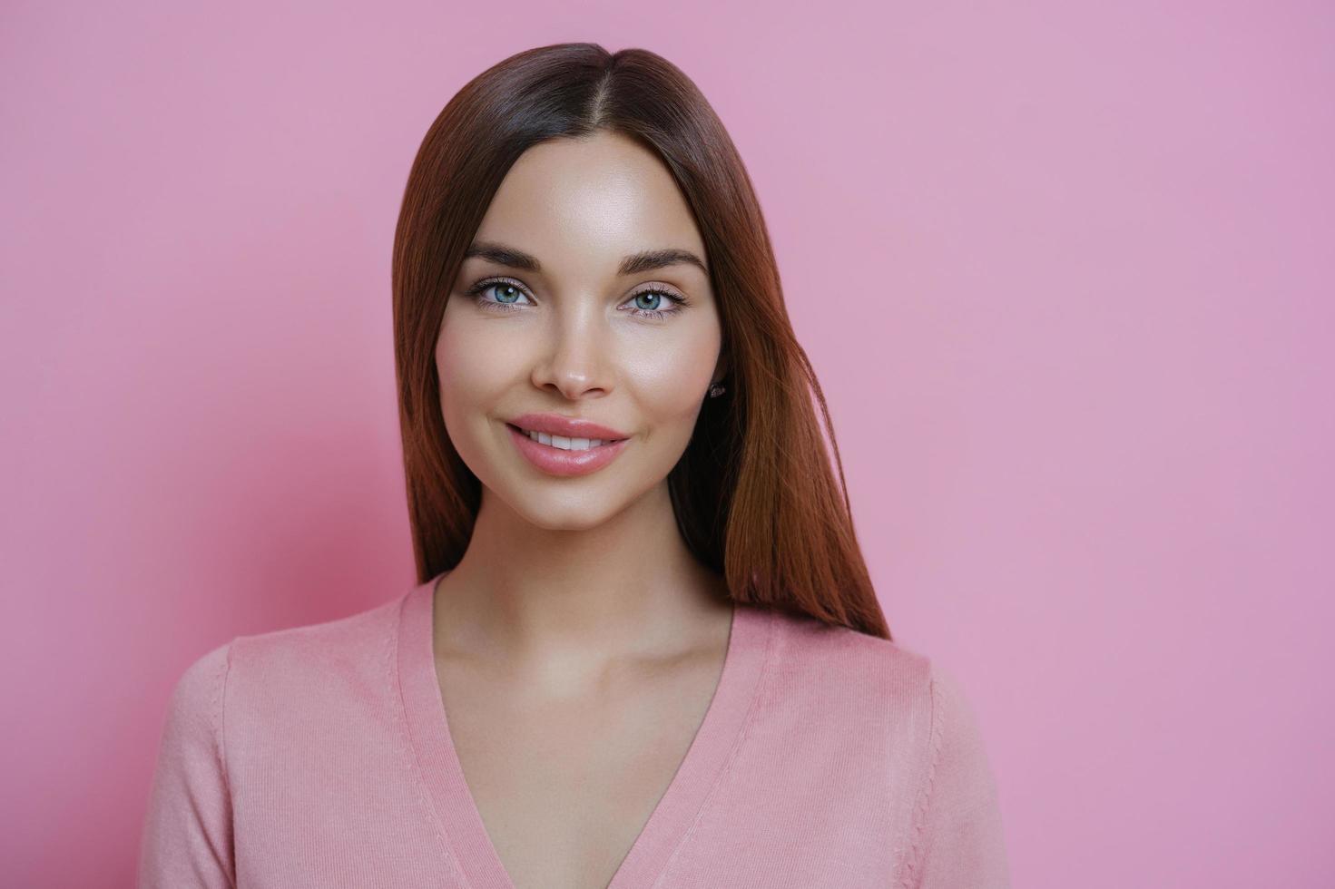 Close up portrait of attractive dark haired woman with brilliant smile, stands indoor against rosy background, has appealing look, isolated over pink background. Face model. Perfect fresh skin photo