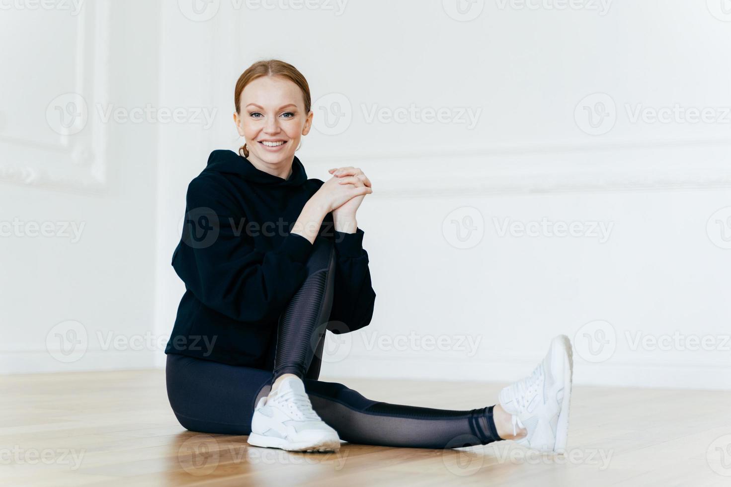 People, sport, recreation concept. Delighted red haired female has positive smile, sits on floor in empty room, leads healthy lifestyle, does fitness exercises, wears hoody, leggings and sneakers photo