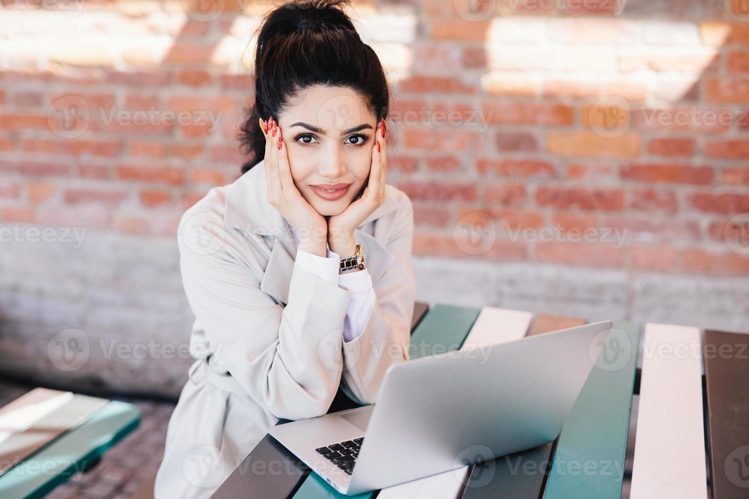 Lovely young brunette woman with charming face and red manicure wearing white coat sitting at table using laptop computer chatting with her family using free internet connection with high speed photo