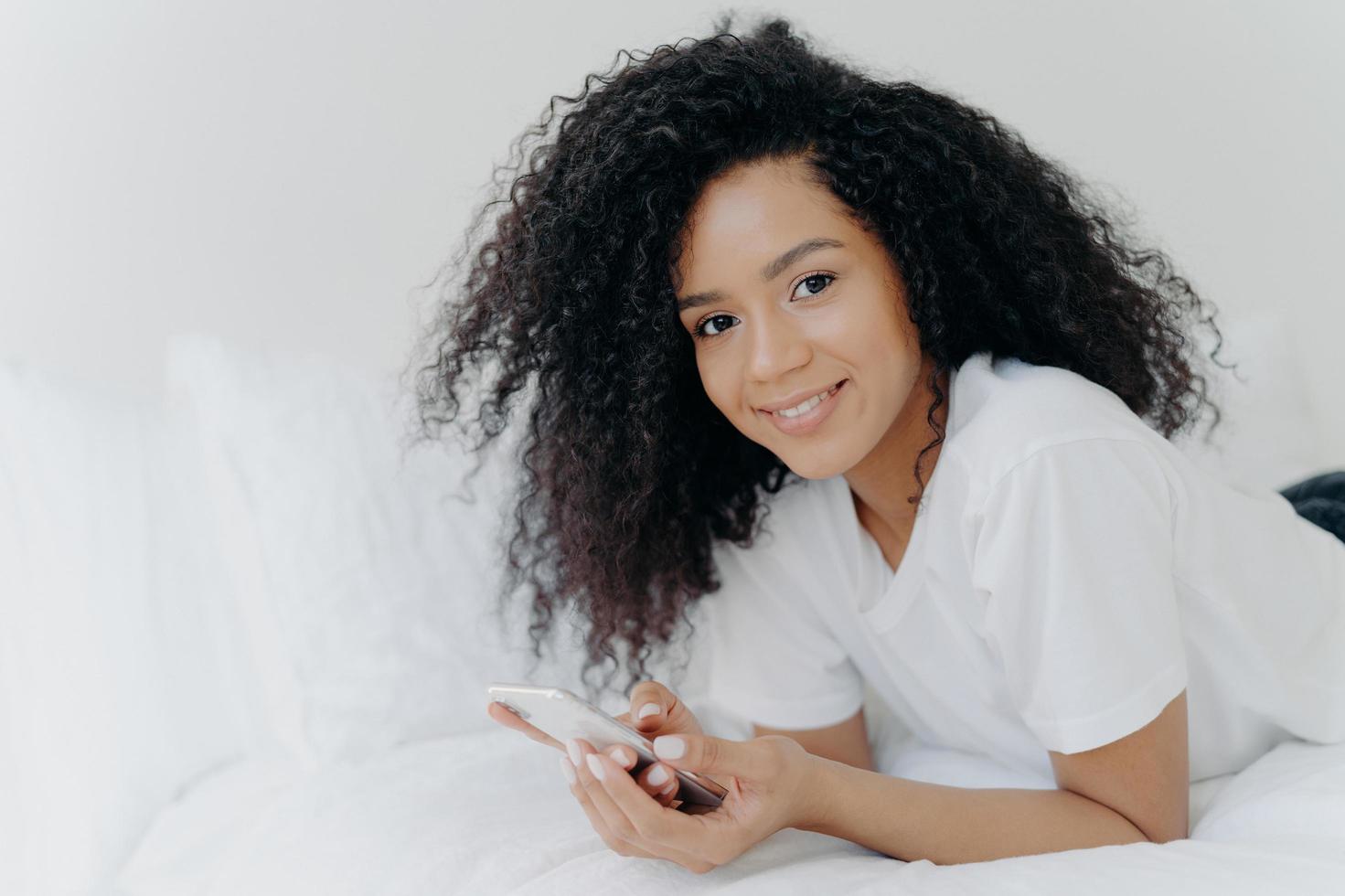 Shot of lovely curly woman with dark skin, tender smile on face, lies on white bed, wears t shirt, has relax with modern technologies, poses in bedroom, reads text message, uses free internet photo