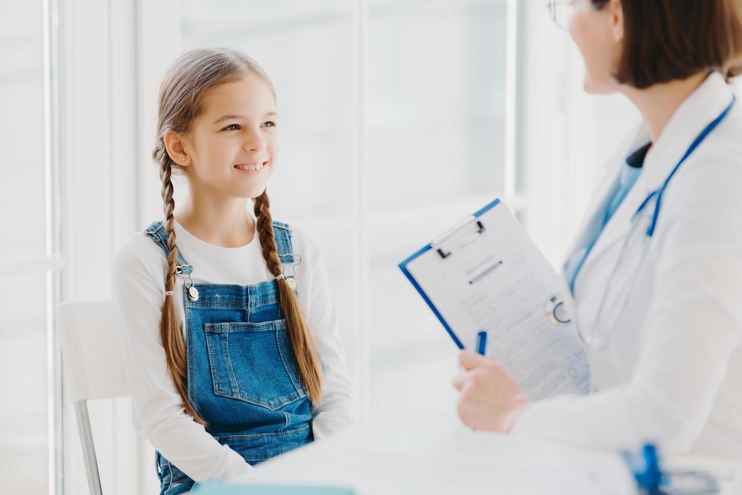 Happy little child listens attentively doctors advice and prescription, comes to see pediatrician in clinic, has two pigtails, wears denim overalls, talk about health problems, has checkup examination photo