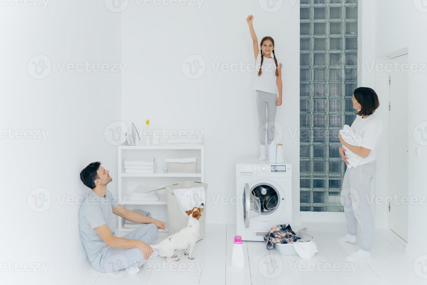Photo of cheerful girl stands on washing machine, raises arm with clenched fist, helps parents to wash clothes, mother, father and dog look at kid. Family in laundry room, busy with domestic chores