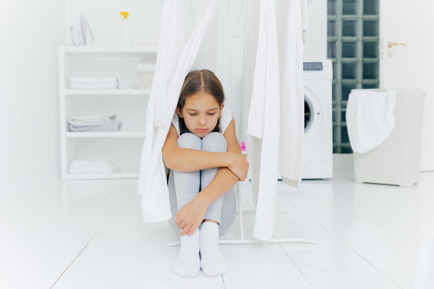 pequeña niña adorable sentada en el piso, siendo castigada por los padres, posa cerca de la secadora de ropa, enfocada con expresión triste, lavadora, canasta con ropa y consola, piensa en algo foto