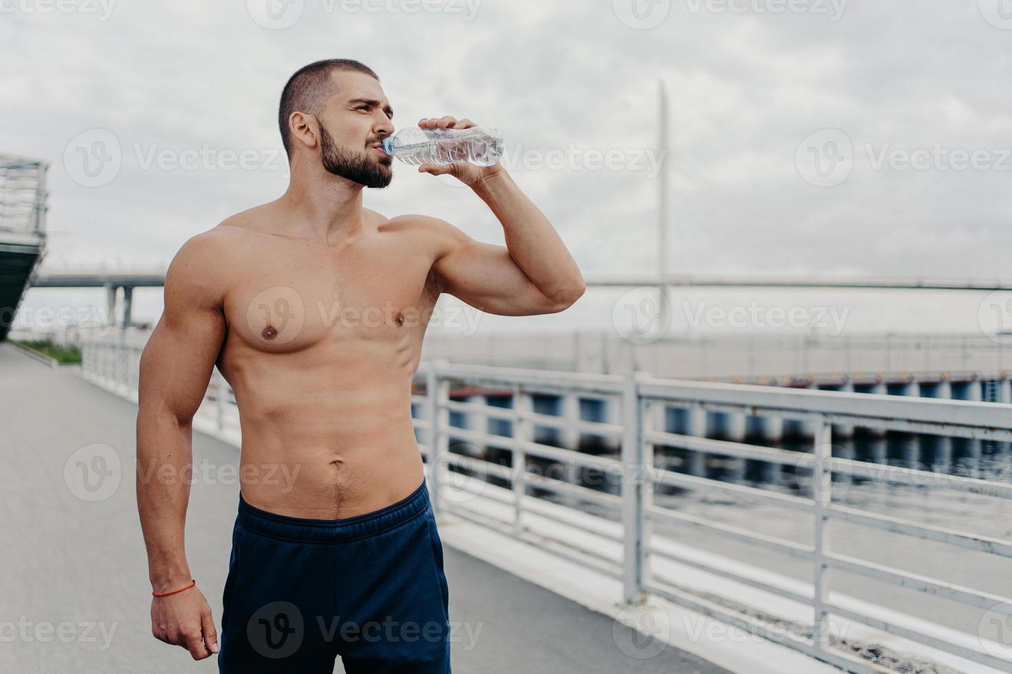 Active sportsman drinks bottle of water poses with naked torso concentrated into distance feels thirsty after running outdoors being full of energy, leads active lifestyle. Sport for better life photo