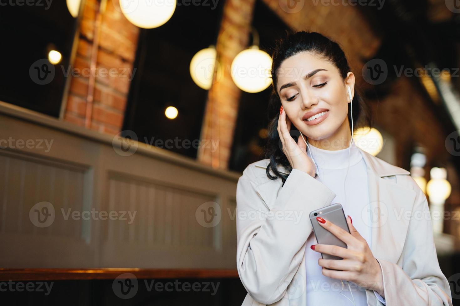 Portrait of beautiful female with dark hair tied in pony tail dressed formally listening to music with earphones closing her eyes with relaxation holding cell phone. People, lifestyle, technology photo