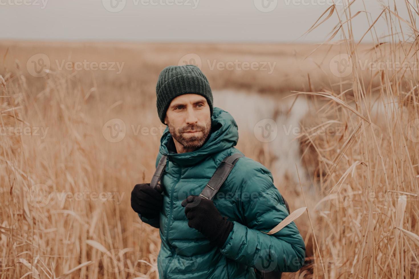 Shot of handsome young man with dark stubble, carries backpack, looks back, notices something, has thoughtful expression, spends free time in open air. Autumn time. People and adventure concept photo