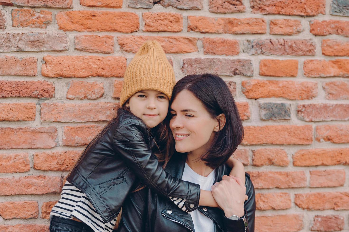 mujer morena sonriente y su pequeña hija se abrazan con amor, tienen sonrisas encantadoras, usan chaquetas de cuero negro, posan contra la pared de ladrillo. la joven madre morena pasa su tiempo libre con una niña foto