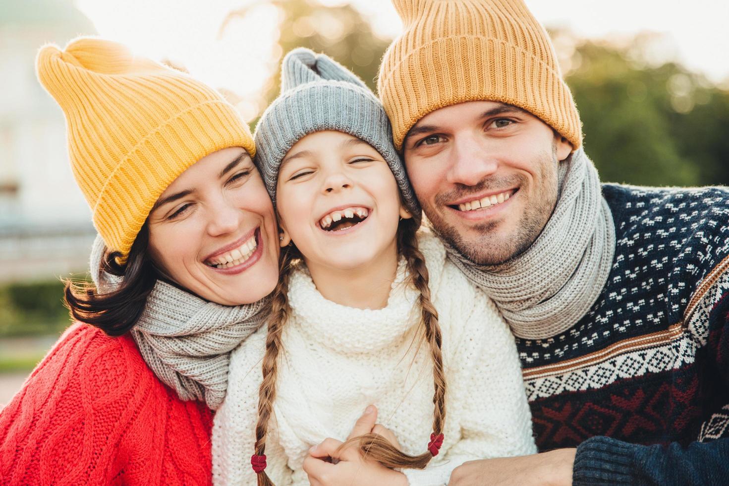 Happy girl with pigtails, wears warm knitted sweater, stands between father and mother, laugh happilly, have sincere smiles on their faces. Relaxed family have holidays, spend nice time together photo