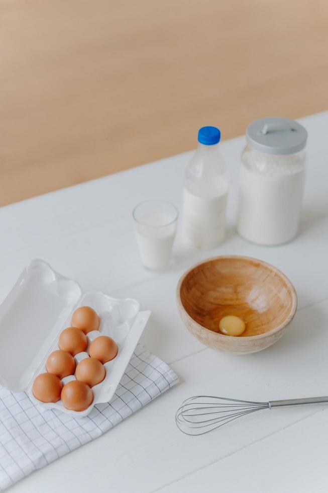 Ingredients for making dough or pasrty. Fresh eggs, milk and flour with whisk on white wooden kitchen table. Products for cooking. Vertical shot photo