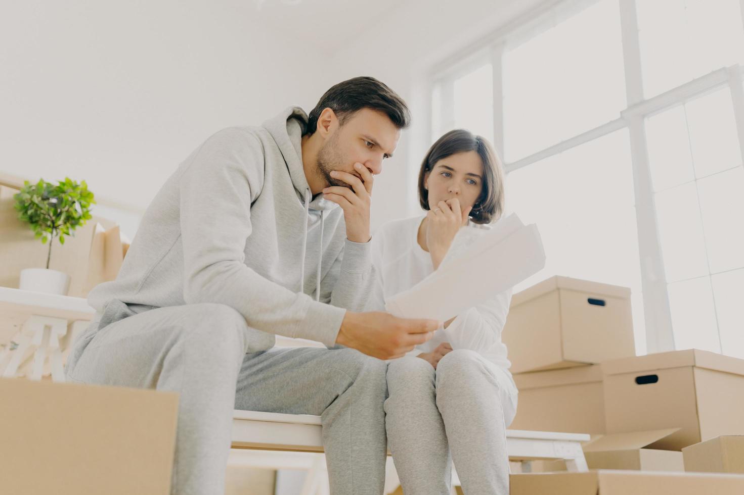 Upset couple stressed by bad news, receive high taxes, looks attentively at papers, read paper letter, have to pay much for flat, move in other apartment, pack belongings in cardboard containers photo