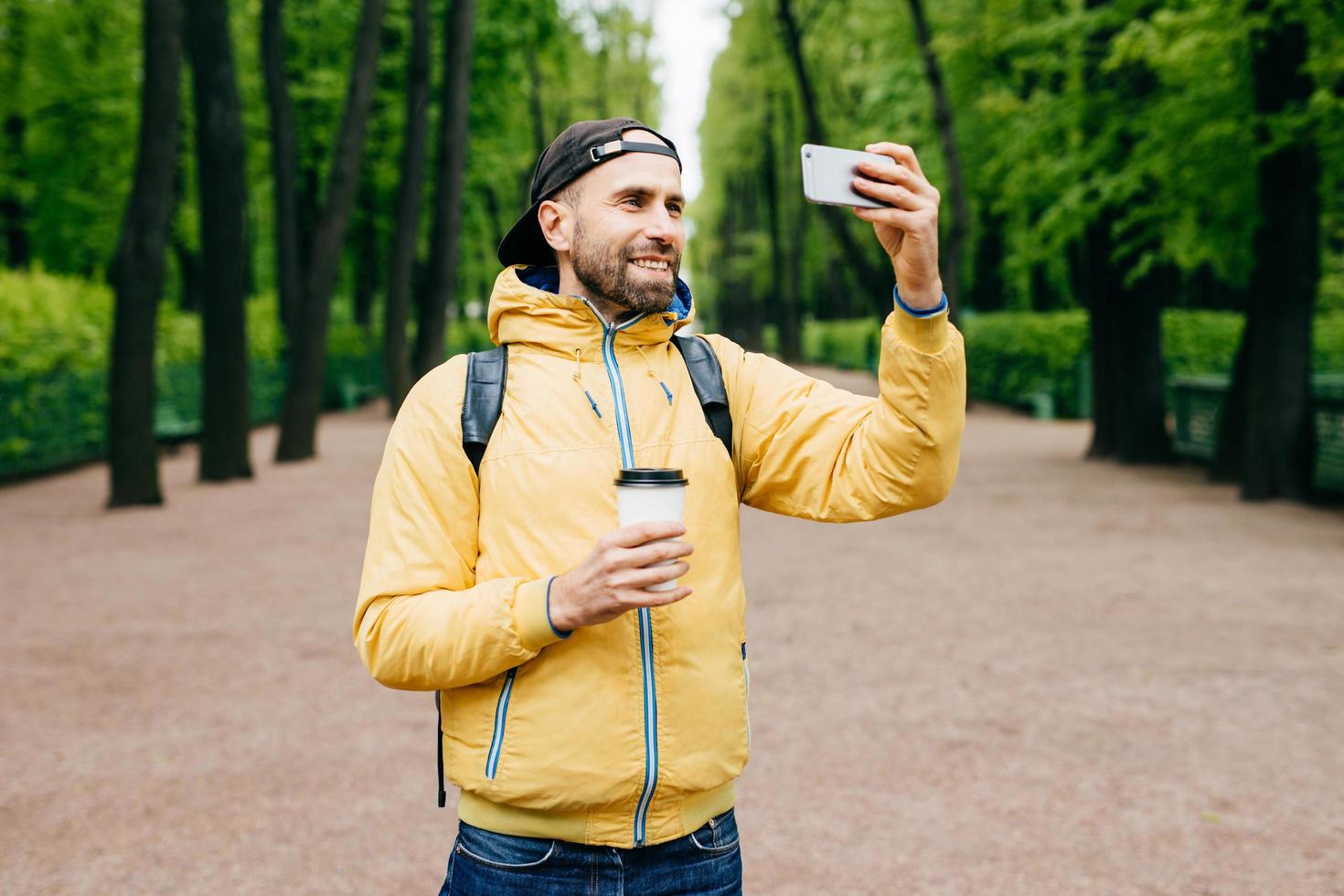 Outdoor portrait of stylish man with stubble beard wearing yellow anorak and holding backpack and takeaway coffee making selfie with his cell phone while standing in beautiful green park having smile photo