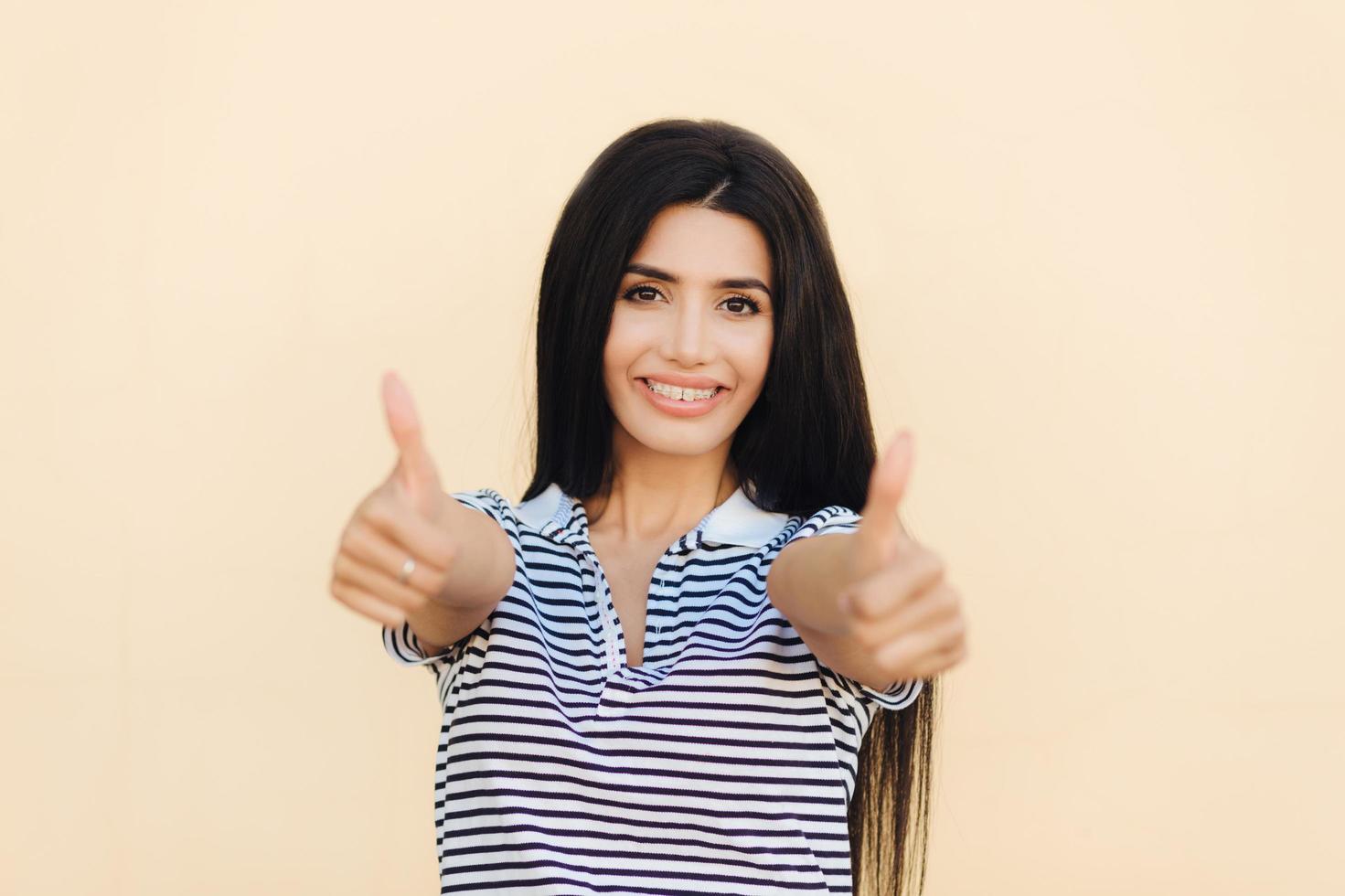 Success and approval concept. Positive female with black long hair, has pleasant smile, keeps thumbs up, likes something, isolated over beige background. Woman celebrates her victory and achievements photo