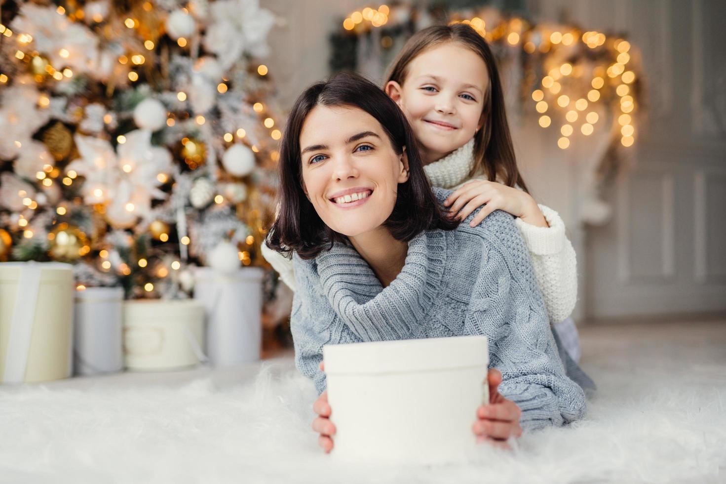 Indoor shot of mother and daughter have fun together, share presents, being in room decorated with garlands and Christmas tree, have joyful expressions, enjoy weekends and winter holidays photo