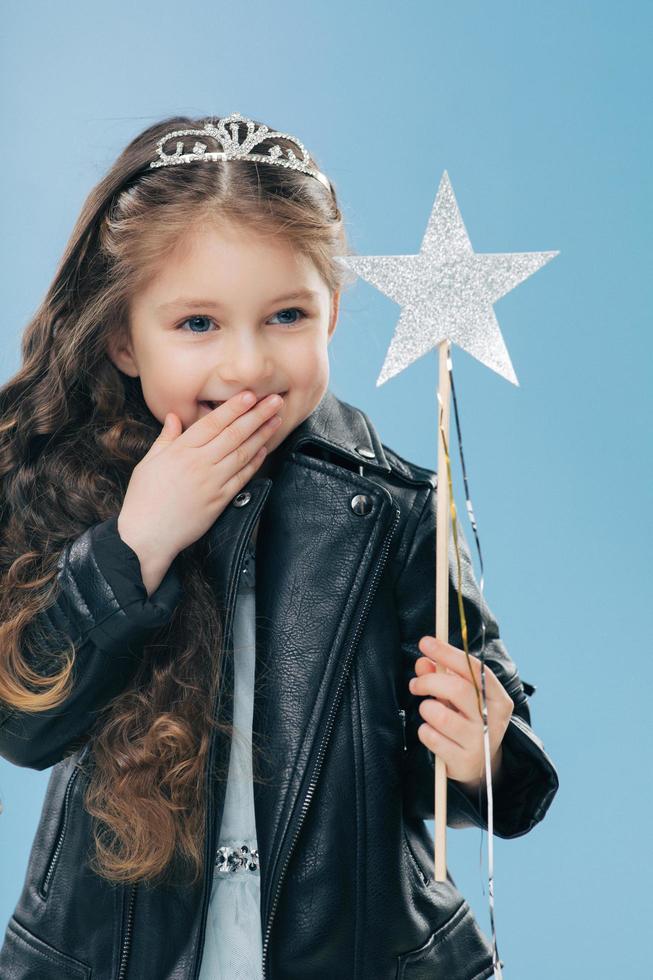 Glad pleasant looking small kid covers mouth with palm, giggles positively, wears crown and black leather jacket, holds magic wand, isolated over blue background. Children and happiness concept photo