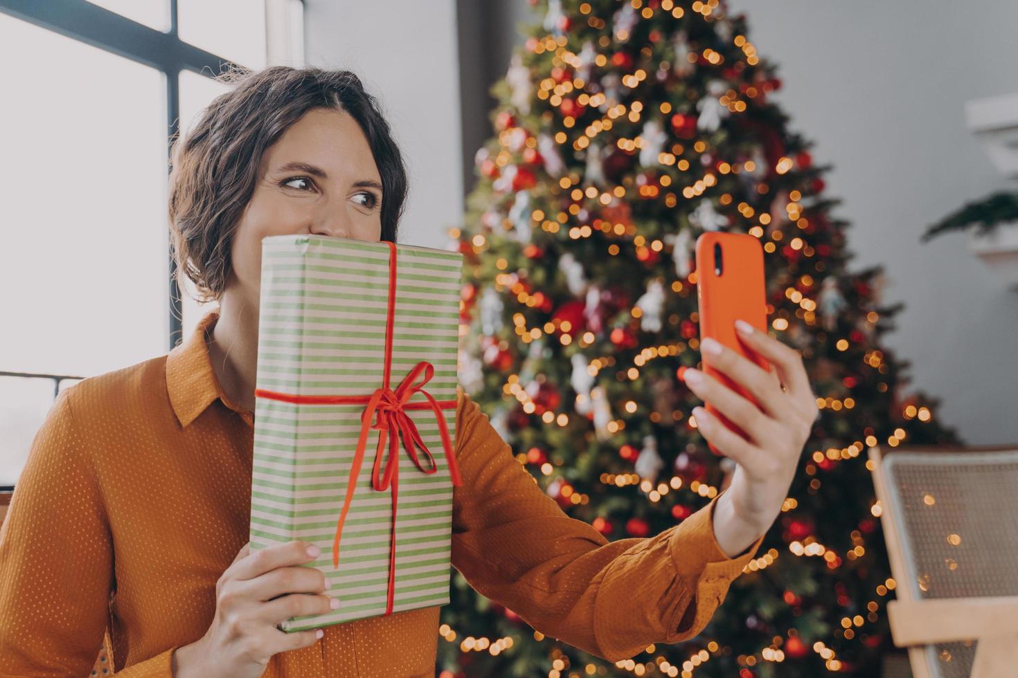 Joyful Spanish bright brunette lady making selfie on smartphone while posing with Xmas gift photo